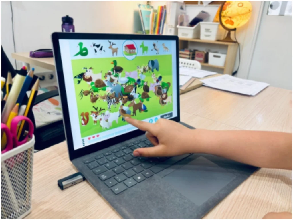 A tablet with a colorful NeuronUP interface and animals, a child’s hand pointing at the screen, and pencils and a balloon on the table.