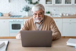 Older man sitting in front of a laptop in a modern kitchen, smiling; stove, white brick wall, and plants in the background.