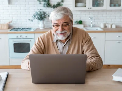 Older man sitting in front of a laptop in a modern kitchen, smiling; stove, white brick wall, and plants in the background.