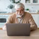 Older man sitting in front of a laptop in a modern kitchen, smiling; stove, white brick wall, and plants in the background.