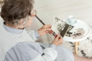 Older woman sitting and looking at a black-and-white photograph; she wears glasses, has gray hair, and uses a cane; more photos and a cup are on the table.