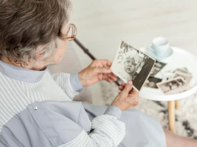 Older woman sitting and looking at a black-and-white photograph; she wears glasses, has gray hair, and uses a cane; more photos and a cup are on the table.