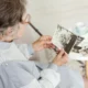 Older woman sitting and looking at a black-and-white photograph; she wears glasses, has gray hair, and uses a cane; more photos and a cup are on the table.