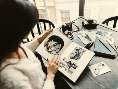 Person sitting at a table looking at a black-and-white photo album, surrounded by notebooks, loose photographs, and a camera.