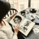 Person sitting at a table looking at a black-and-white photo album, surrounded by notebooks, loose photographs, and a camera.